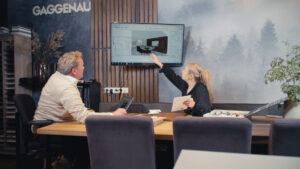 Twee mensen zijn aan het werk. Ze zitten aan een grote tafel en kijken naar een tv waarop ze een keuken aan het ontwerpen zijn.