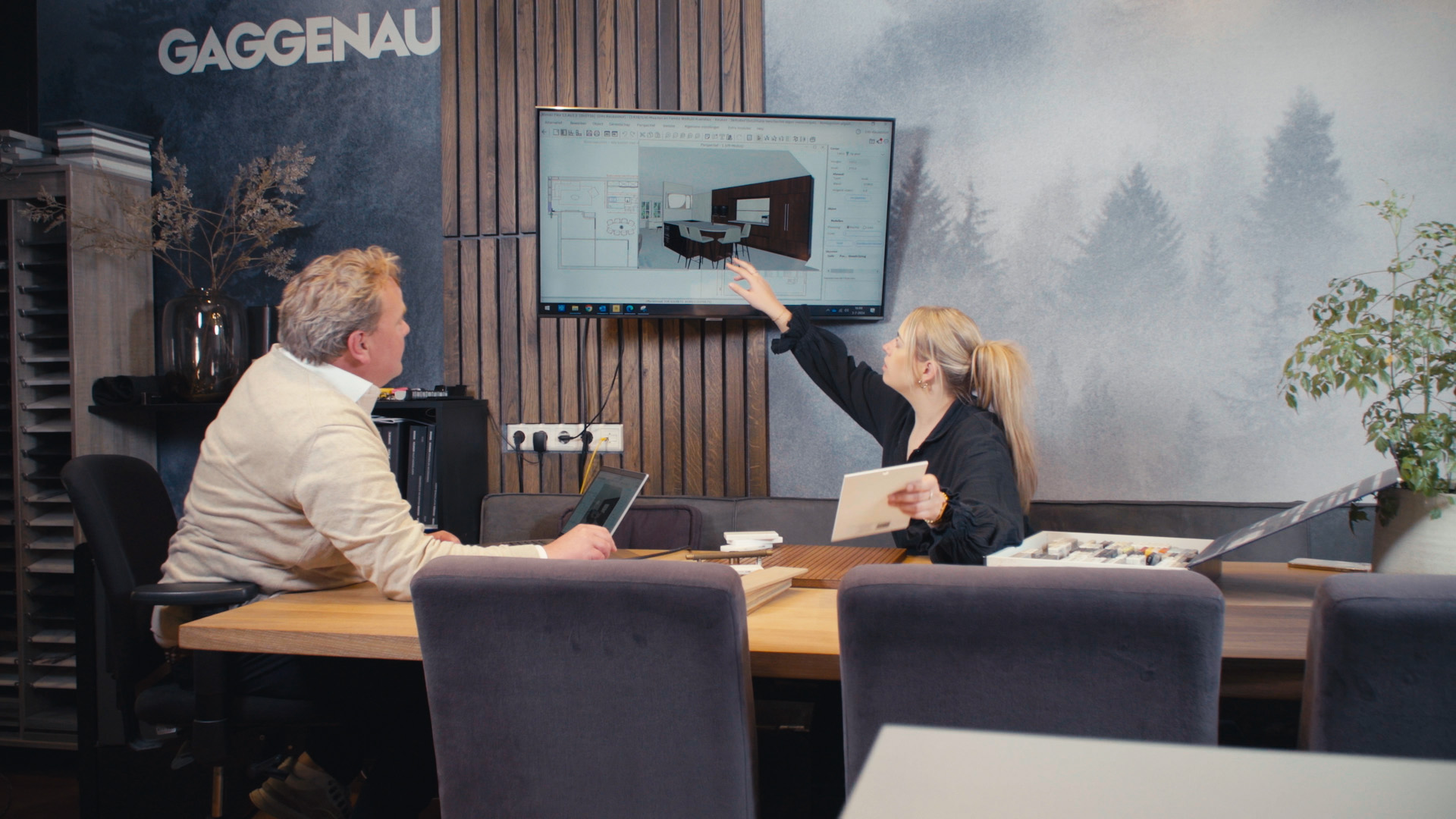 Twee mensen zijn aan het werk. Ze zitten aan een grote tafel en kijken naar een tv waarop ze een keuken aan het ontwerpen zijn.