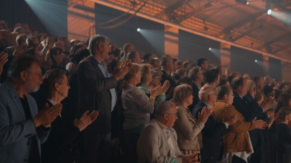 Een groot publiek in een zaal kijkt naar rechts en geeft een staand applaus